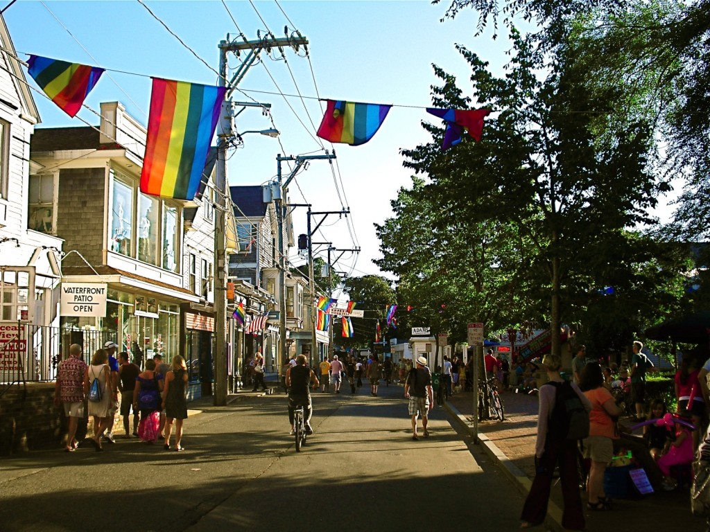 Rainbow flags flutter over shop lined street with many pedestrians 