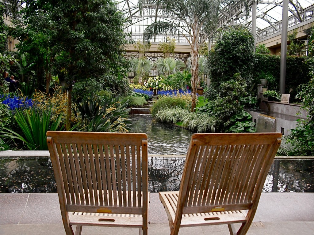 Teak chairs face landscaped pond within glass conservatory building. Conservatory, Longwood Gardens, PA #visitphilly #PATravelHappy @GetawayMavens
