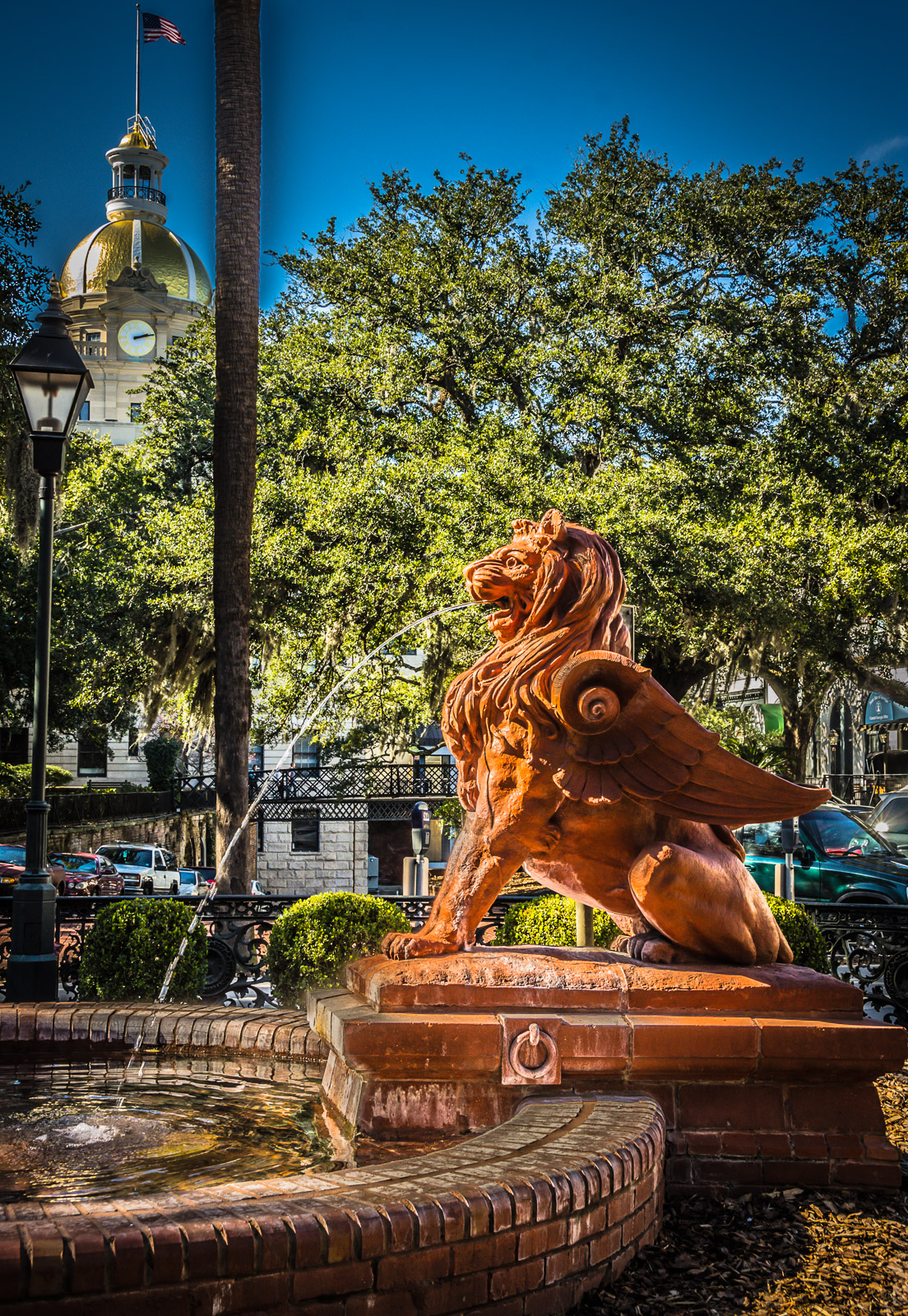 Savannah GA's spouting Lion Fountain in front of the Cotton Exchange, with gold roof of Savannah City Hall peeking out amidst trees in the background.