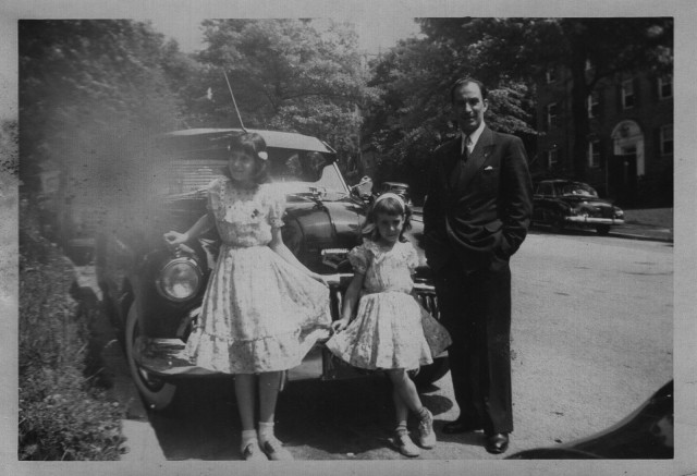 Two girls in frilly dresses with their father in front of a big, black Buick in the early 1950s.