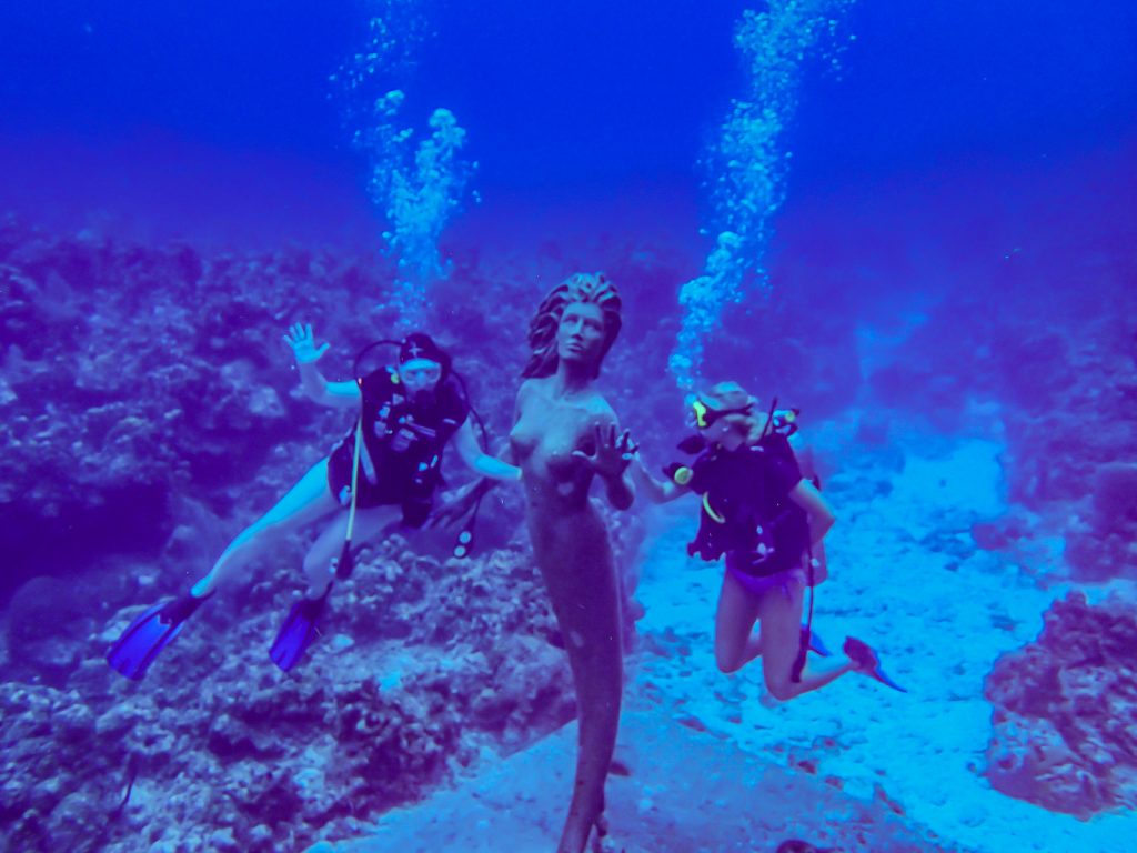 Two scuba divers pose underwater with the Mermaid Statue in the Grand Cayman