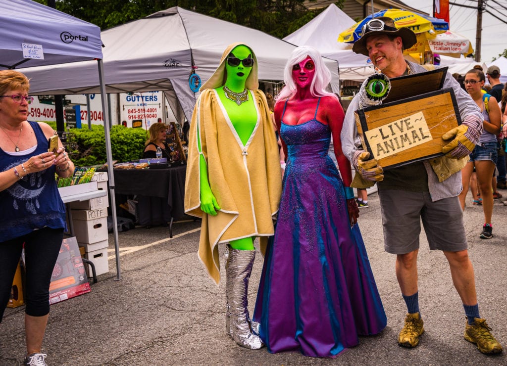 A man dressed in a safari hunter costume carries a box labelled Live Animal that has a one-eyed extraterrestrial popping out. Next to him, two women dressed as aliens pose with him as a tourist captures the image on an iPhone.