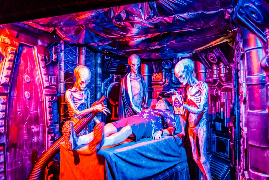 Tourist lies on examining table where he is surrounded by aliens at UFO Fair pop-up museum.