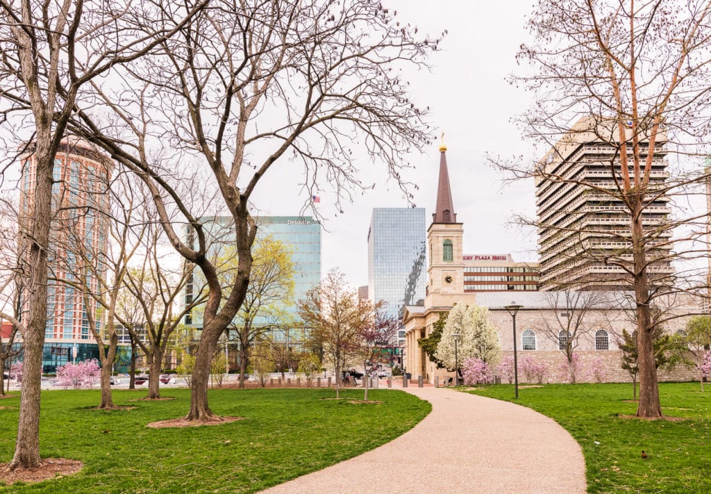 Curved walkway lined with trees covered in Spring blooms leading up to Old Cathedral with modern buildings of Downtown St Louis in background.