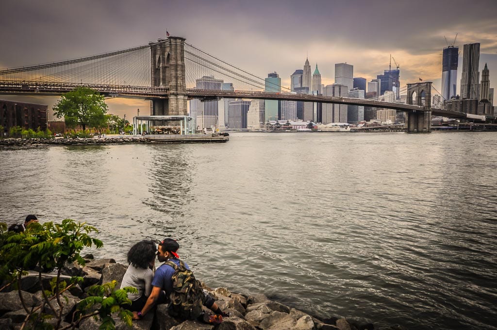 Couple smooches in front of sunset view of the Brooklyn Bridge.