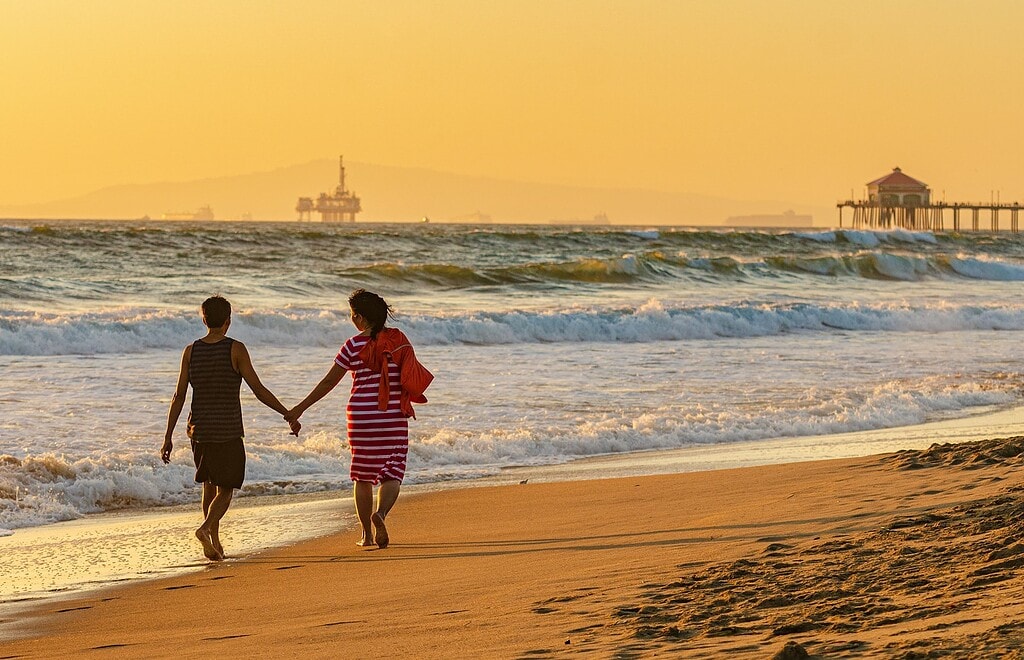 Couple strolls holding hands on Huntington Beach in California. This is an example of why couples should travel together.